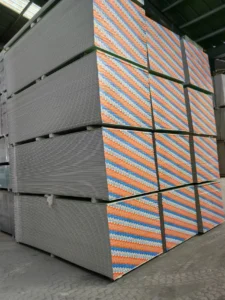 Large stack of drywall sheets with colorful patterned edges in a warehouse setting, under a corrugated metal roof.