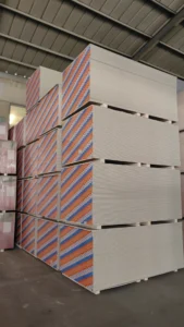 Stacks of drywall sheets with colorful edges in a warehouse setting, seen from below the metal ceiling.