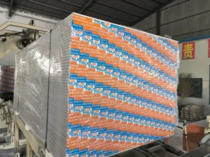 A large stack of gypsum boards wrapped in plastic, labeled 'Trusus', in a manufacturing or storage facility.