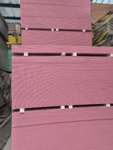 Stacks of pink drywall sheets organized in a warehouse with metal roofing visible above and other materials around.