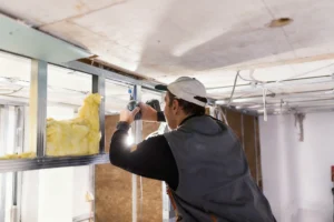 A construction worker using a drill to install insulation in a metal framework for ceiling construction, wearing a cap and work vest.