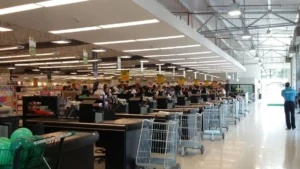 Supermarket checkout area with multiple counters, shopping carts, and customers waiting in line under bright lighting.