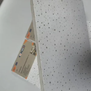 Perforated ceiling tiles with visible holes, stacked together. Light gray color with rough edges and packaging visible in the background.