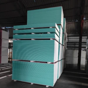 Large stacks of green and white drywall panels in an industrial warehouse setting under a high ceiling.