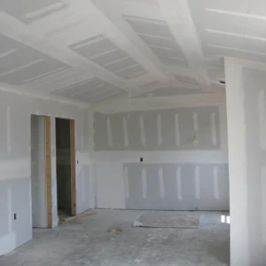 Room under construction with freshly installed drywall and ceiling panels, featuring exposed door frames and electrical wiring.